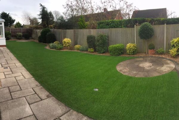 A well-maintained backyard with neatly cut artificial grass, bordered by a stone pathway. Various shrubs and plants line a wooden fence, framing a circular paved patio area in the center, surrounded by more greenery. The sight raises questions about how such lush beauty is achieved amidst visible houses in the background.